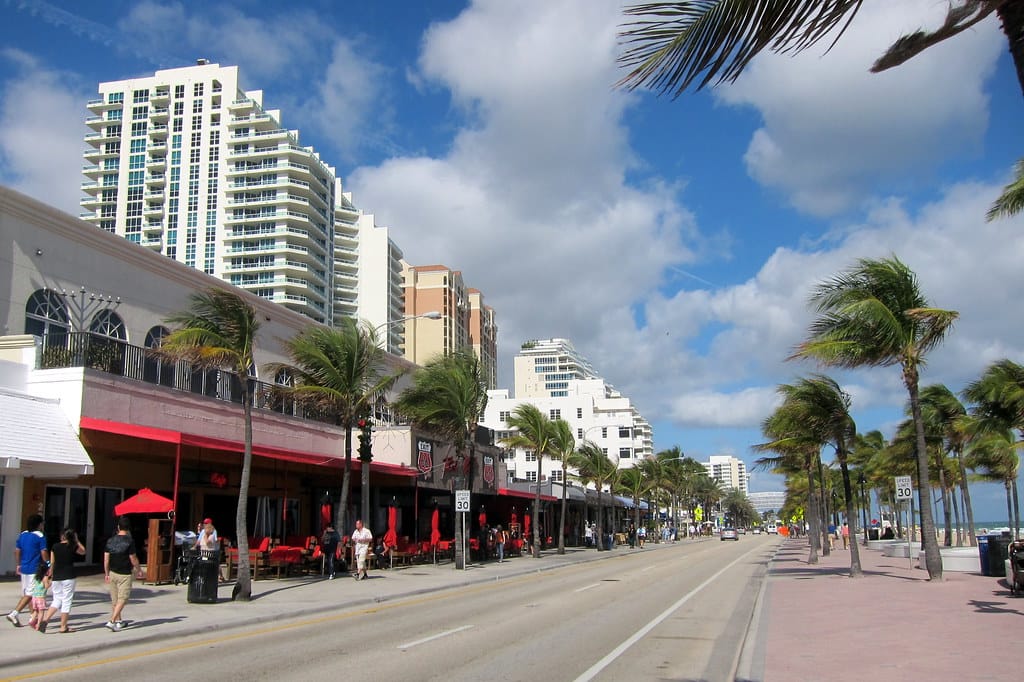 Palm-lined beachfront promenade along A1A in Fort Lauderdale, with people walking beside shops and towers